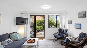 Ein Wohnzimmer mit einer Couch, zwei Korbstühlen, einem Couchtisch, einem Fernseher und einer Schiebetür zu einem Garten.