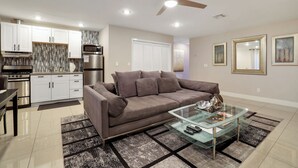 A modern living room with a glass coffee table, a sofa, a ceiling fan, and a kitchen area with white cabinets and stainless steel appliances. - Perfect Group Getaway w/ Private Pool (Las Vegas)