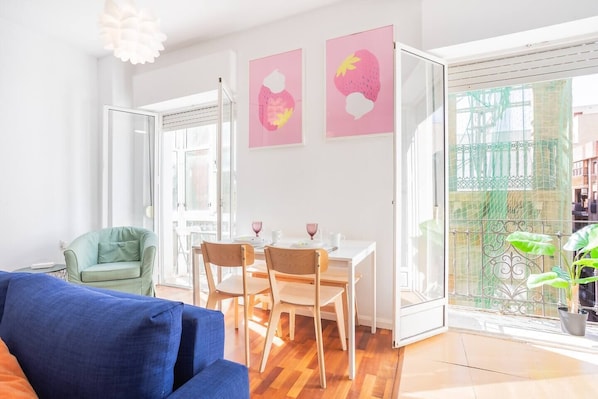 Un salon lumineux avec un canapé bleu, une table à manger en bois et des chaises, un fauteuil vert et deux œuvres d’art murales roses.