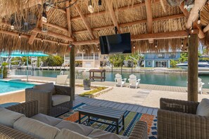 A covered outdoor area with wicker furniture, a pool, and a TV screen. - Waterfront Pool Home w/ 100' Dock, Tiki Hut & Boating Access Near Key West (Sugarloaf Key)