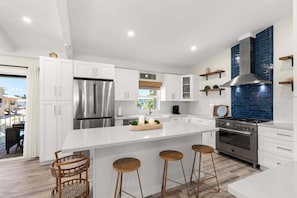 A modern kitchen with white cabinetry, stainless steel appliances, a central island with stools, and a blue-tiled backsplash. - Waterfront Pool Home w/ 100' Dock, Tiki Hut & Boating Access Near Key West (Sugarloaf Key)