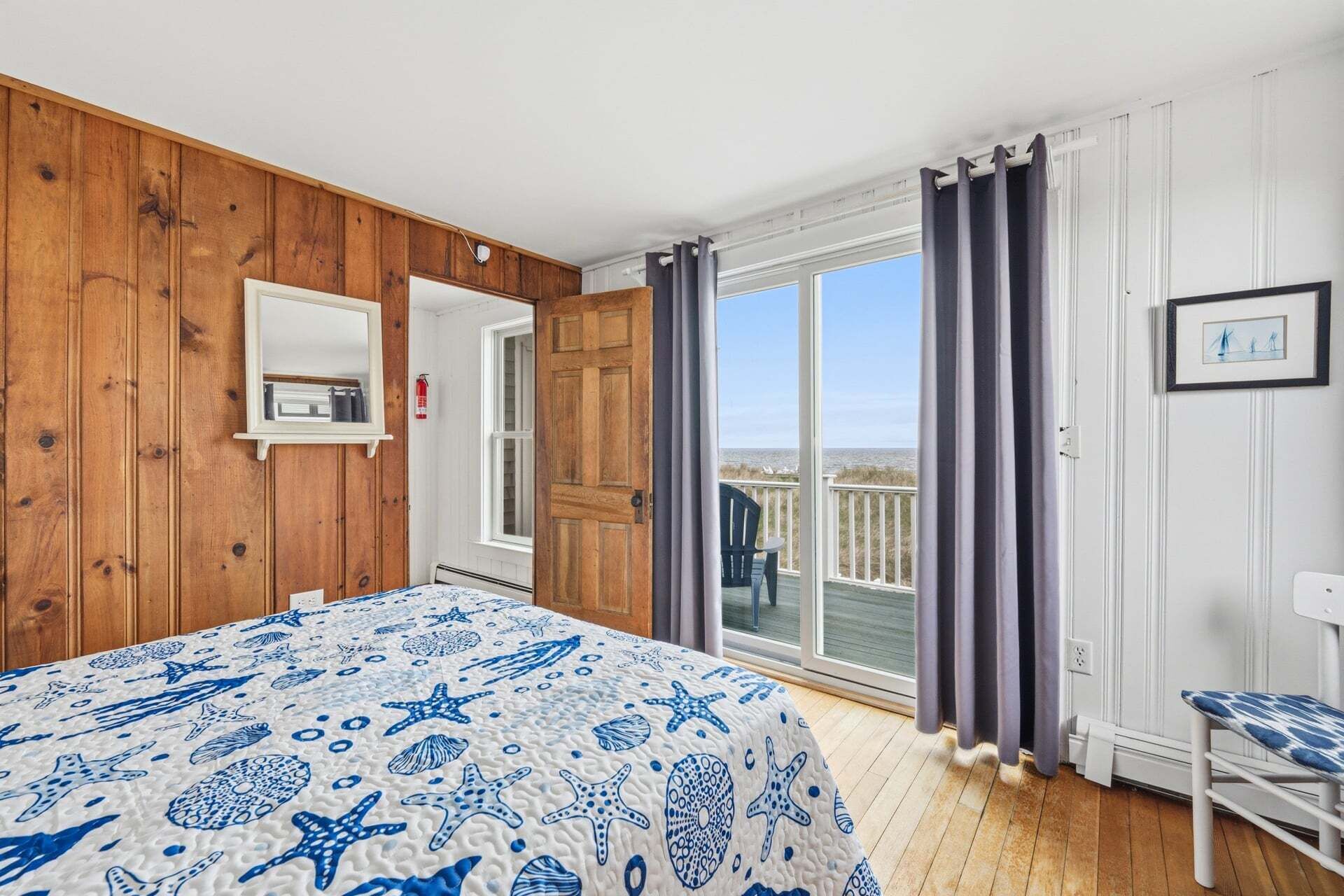 A bedroom with a wooden wall, a bed with a blue and white ocean-themed bedspread, a sliding glass door to a balcony, and a chair.