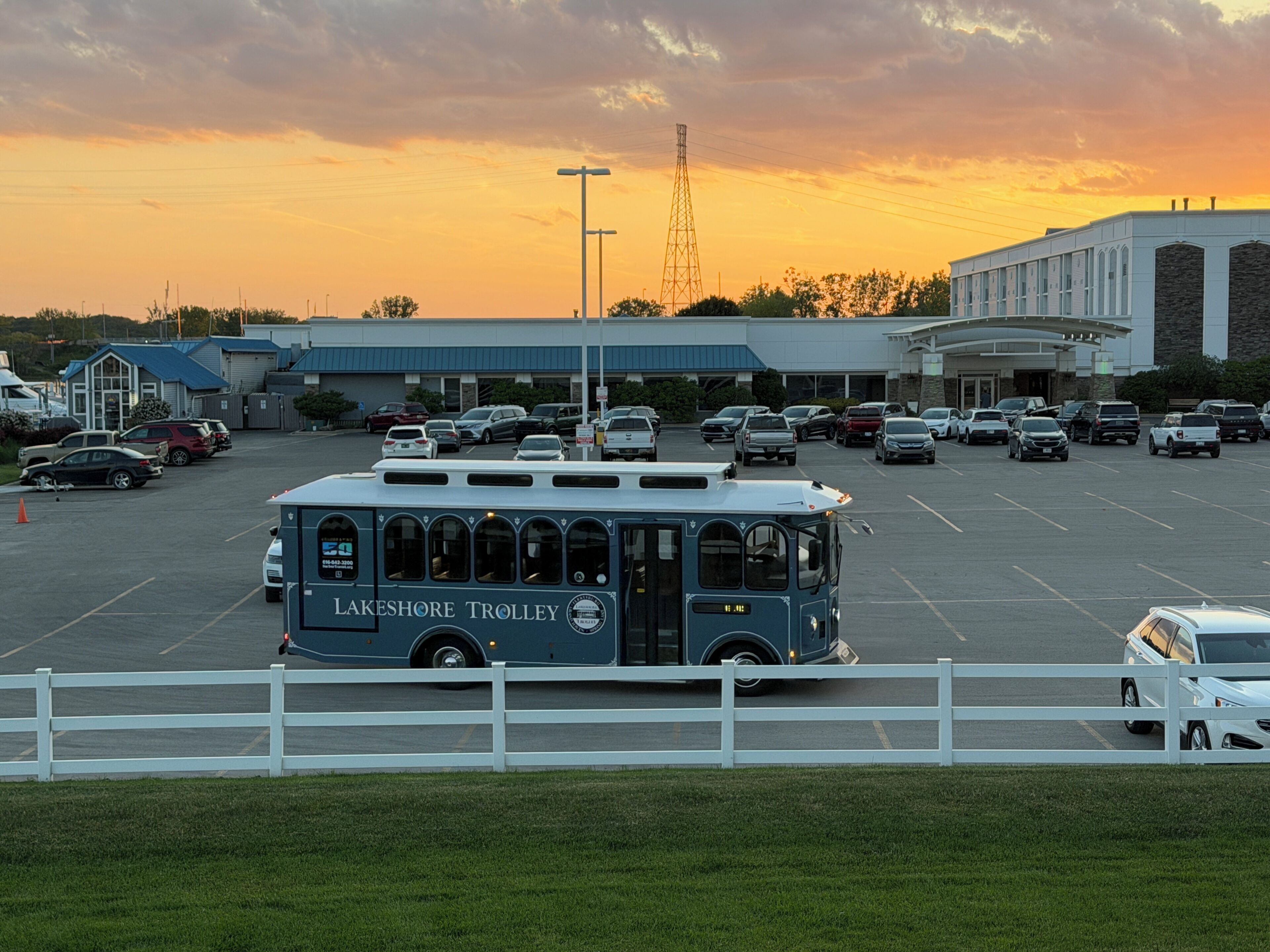 Ein Parkplatz mit einem Trolley mit der Aufschrift 'Lakeshore Trolley', einem Gebäude mit blauem Dach und einem weißen Zaun im Vordergrund.