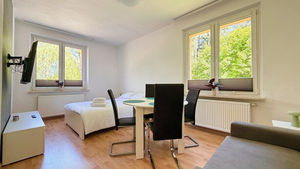 Une pièce comprenant un lit, une table avec des chaises, un radiateur et des fenêtres donnant sur des arbres.