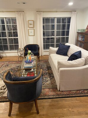 Ein Wohnzimmer mit Sofa, Sessel und Couchtisch, großen Fenstern mit Vorhängen und einem gemusterten Teppich.