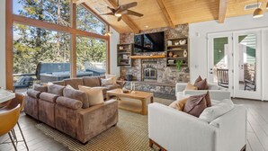 A spacious living room with a stone fireplace, large windows, and a ceiling fan.