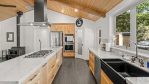A modern kitchen with a central island, stainless steel appliances, wooden cabinetry, and a large window offering a view of the outdoors.