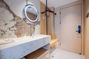 Un bany amb un lavabo de marbre, un mirall rodó amb il·luminació, una banca de fusta i una porta de fusta.
