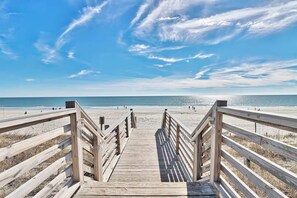 Ein hölzerner Steg führt zu einem Strand bei strahlendem Himmel.