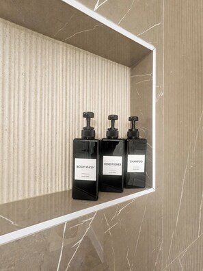 Drei schwarze Flaschen mit Duschgel, Conditioner und Shampoo auf einem Regal vor einer strukturierten Wand.