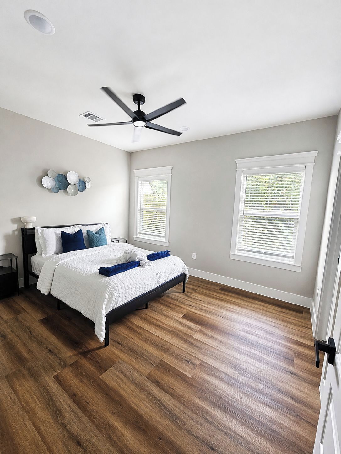 Ein Schlafzimmer mit einem Bett, einem Deckenventilator, zwei Fenstern mit Jalousien und einer Wandleuchte.