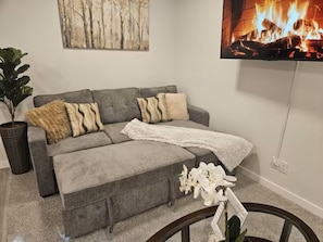 Ein graues Sofa mit vielen Kissen, einer weißen Tagesdecke und einem Bilderrahmen mit einem Feuer.