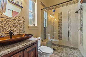 A bathroom with a stone mosaic backsplash, a wooden vanity, a white toilet, and a glass-enclosed shower with mosaic tiles. - 
Unique Multi-Family Getaway with Pool, Game room, Theater & 6 Themed Bedrooms (Spring Branch)