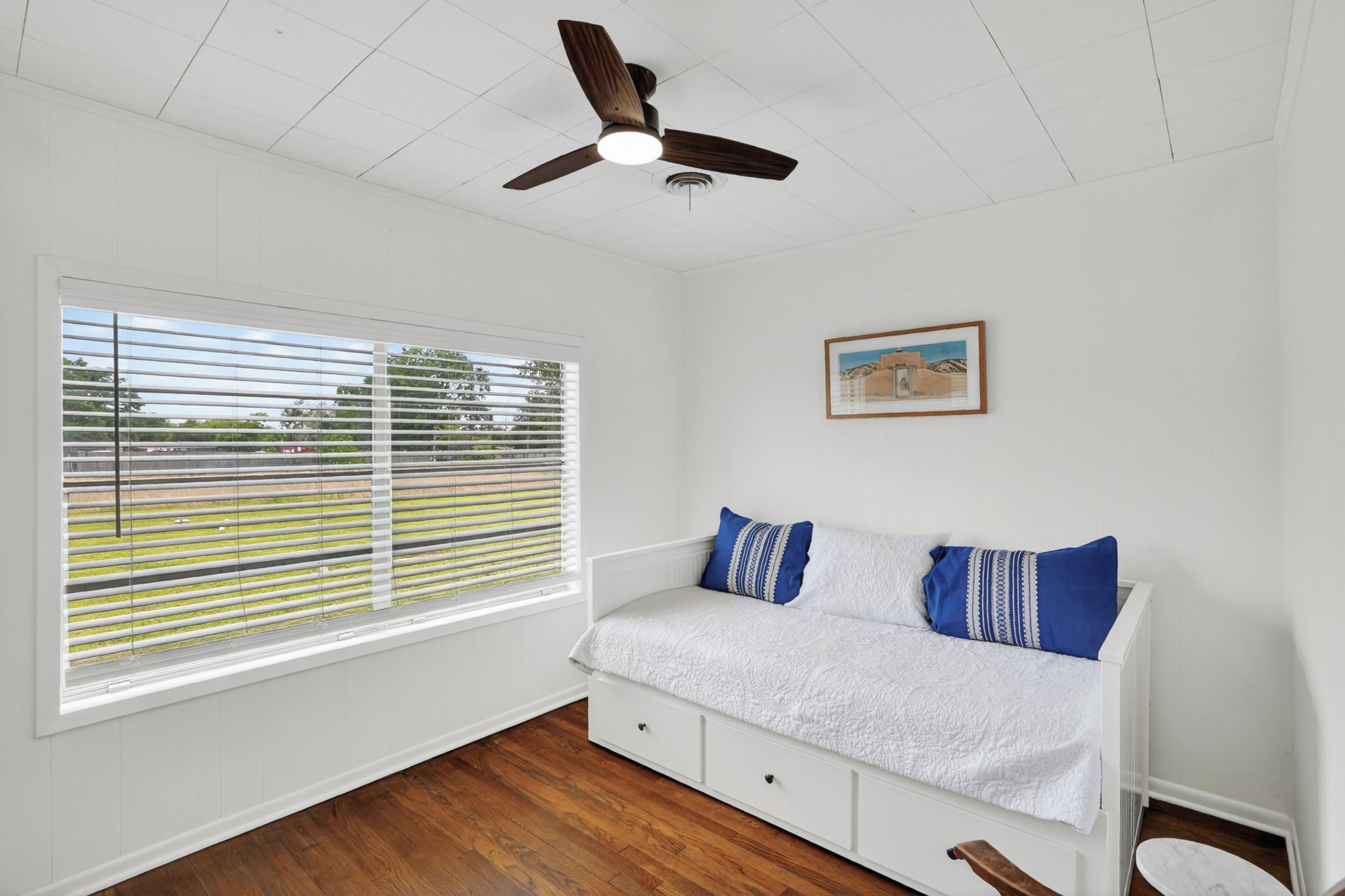 Un dormitorio con cama, ventilador de techo, ventana con persianas e imagen enmarcadas en la pared.