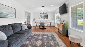 Ein modernes Wohnzimmer mit einem grauen Sofa, einem Essbereich mit rundem Tisch und einem an der Wand montierten Fernseher.