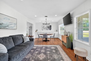 A modern living room with a grey sofa, a dining area with a round table, and a TV mounted on the wall. - The Staytion (College Station)