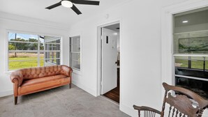 Ein Zimmer mit Deckenventilator, einem Ledersofa, einem Holzstuhl und großen Fenstern mit Blick auf eine grüne Landschaft.
