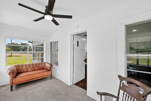 A room with a ceiling fan, a leather sofa, a wooden chair, and large windows with a view of a green landscape. - The Staytion (College Station)