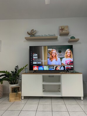 Ein Flachbildfernseher auf einer weißen Unterhaltungsanlage, wobei die Netflix-Oberfläche 'LOOK BOTH WAYS' anzeigt und ein Schild mit der Aufschrift 'Willkommen am Strand' darüber hängt.
