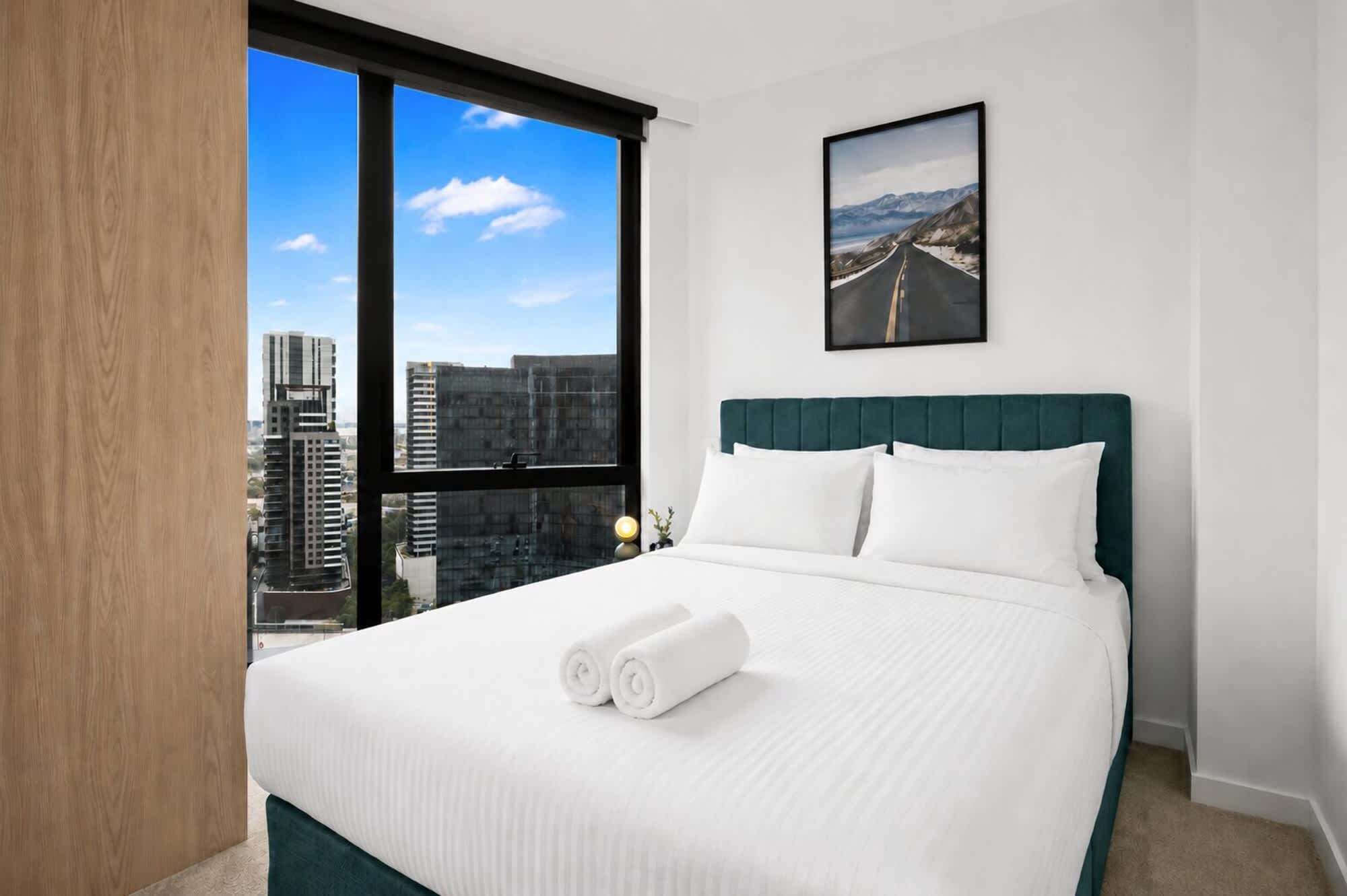 A hotel room with a large window, a bed with white linens, a green headboard, and a framed picture on the wall.