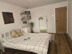 A bedroom with a bed, a bedside table, a lamp, a potted plant, a bookshelf, a mirror, and a wooden door.