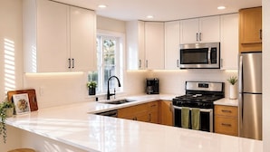 A modern kitchen with white cabinetry, a large island with stools, stainless steel appliances, and a window providing natural light. - Central Ballard Townhome / Sleeps 8 / Direct Bus Line to Stadium (Seattle)