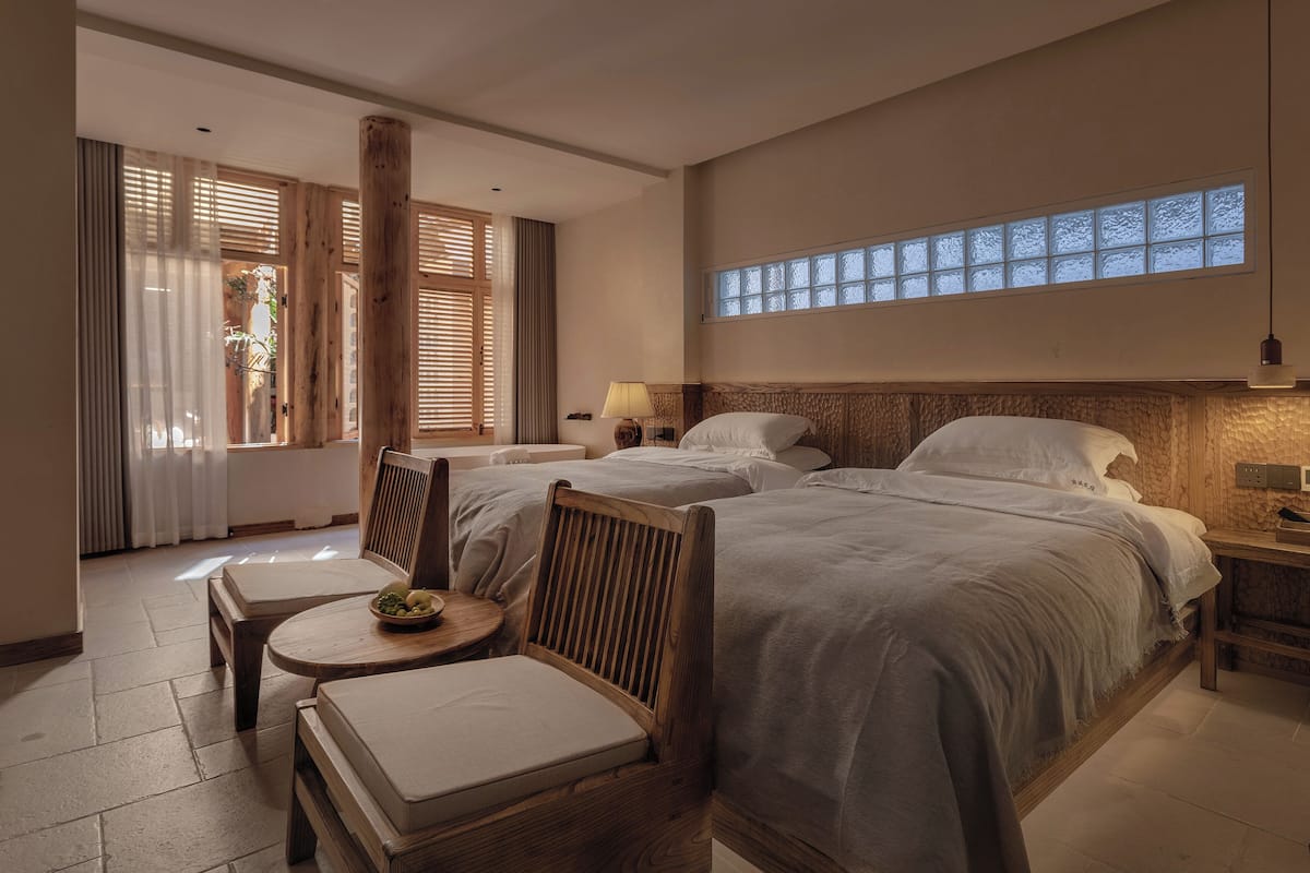 A hotel room with two beds, a small table with a bowl of fruit, and a chair.
