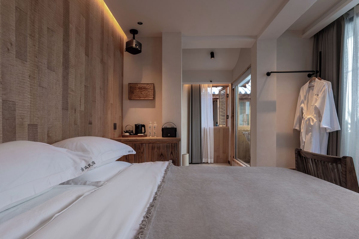 A hotel room with a large bed, a wooden headboard, a bedside table with a kettle, a robe hanging on a rack, and a view through a glass door.