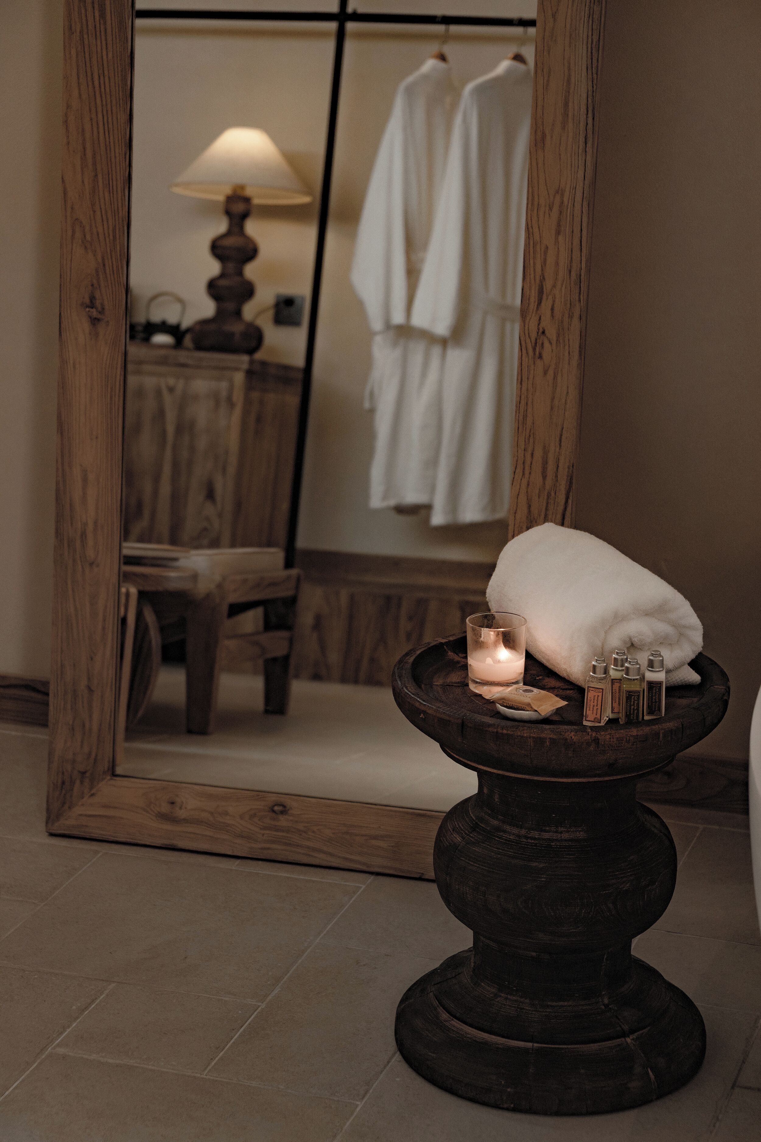 Un miroir à cadre en bois reflète une salle de bain où l’on aperçoit une serviette blanche, une bougie et des articles de toilette posés sur un piédestal.