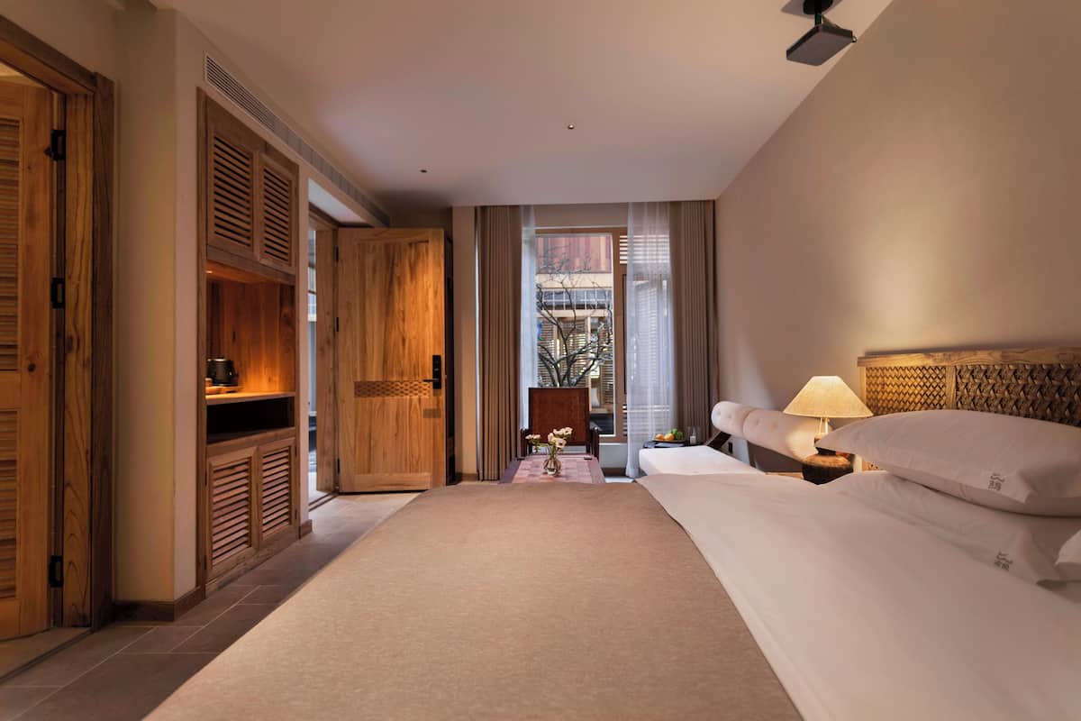 A hotel room with a large bed, a nightstand with a lamp, a wooden wardrobe, and a view of the outside.