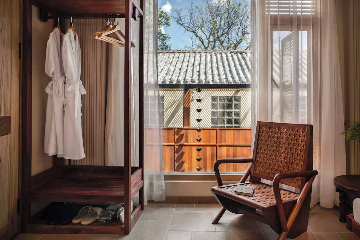 A room with a wooden bench, a chair, a window with a view of a traditional building, and a wooden wardrobe with hanging robes.