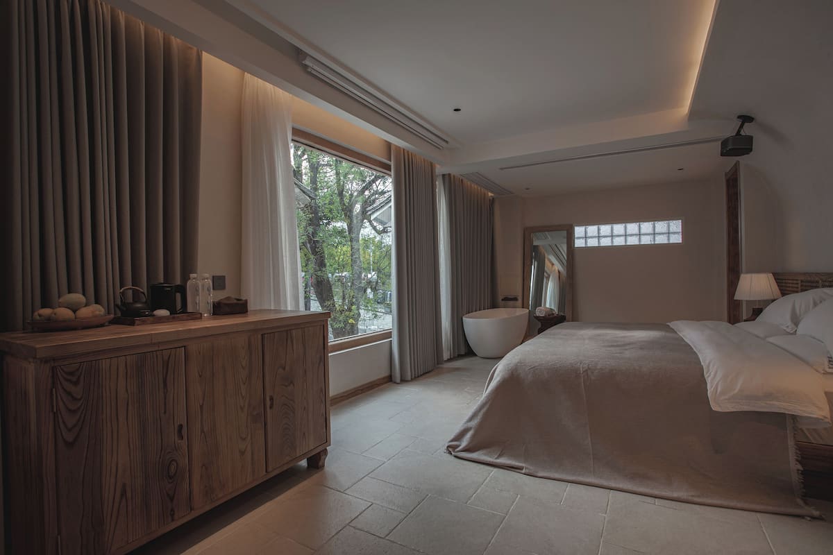 A bedroom with a large bed, a wooden dresser, a bathtub, and a window with curtains.