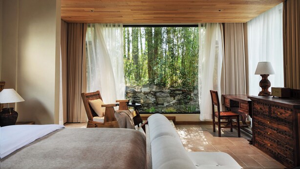 Une chambre d’hôtel avec un lit, deux chaises, un bureau, une lampe et une vue sur une forêt, visible à travers de grandes fenêtres.