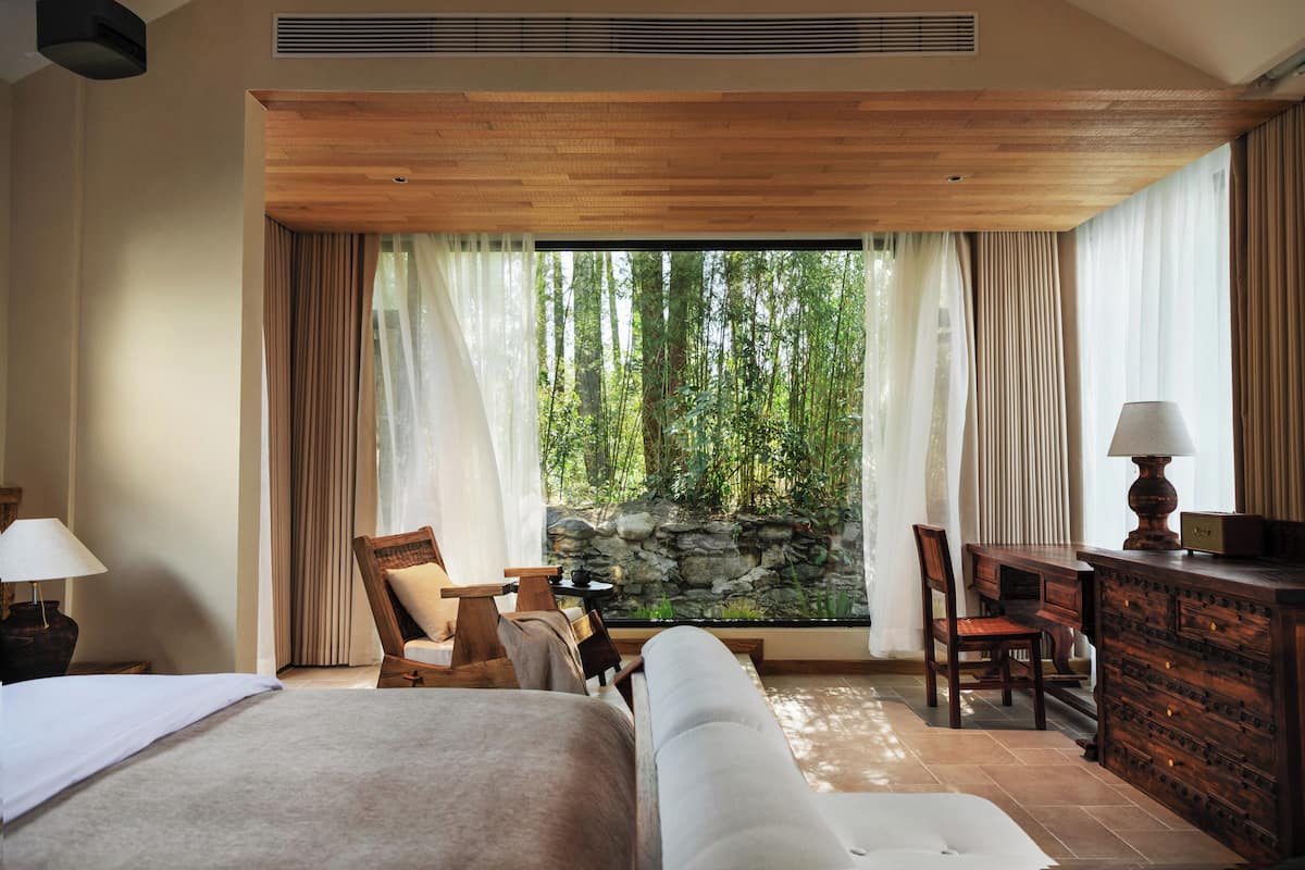 A hotel room with a bed, two chairs, a desk, a lamp, and a view of a forest through large windows.