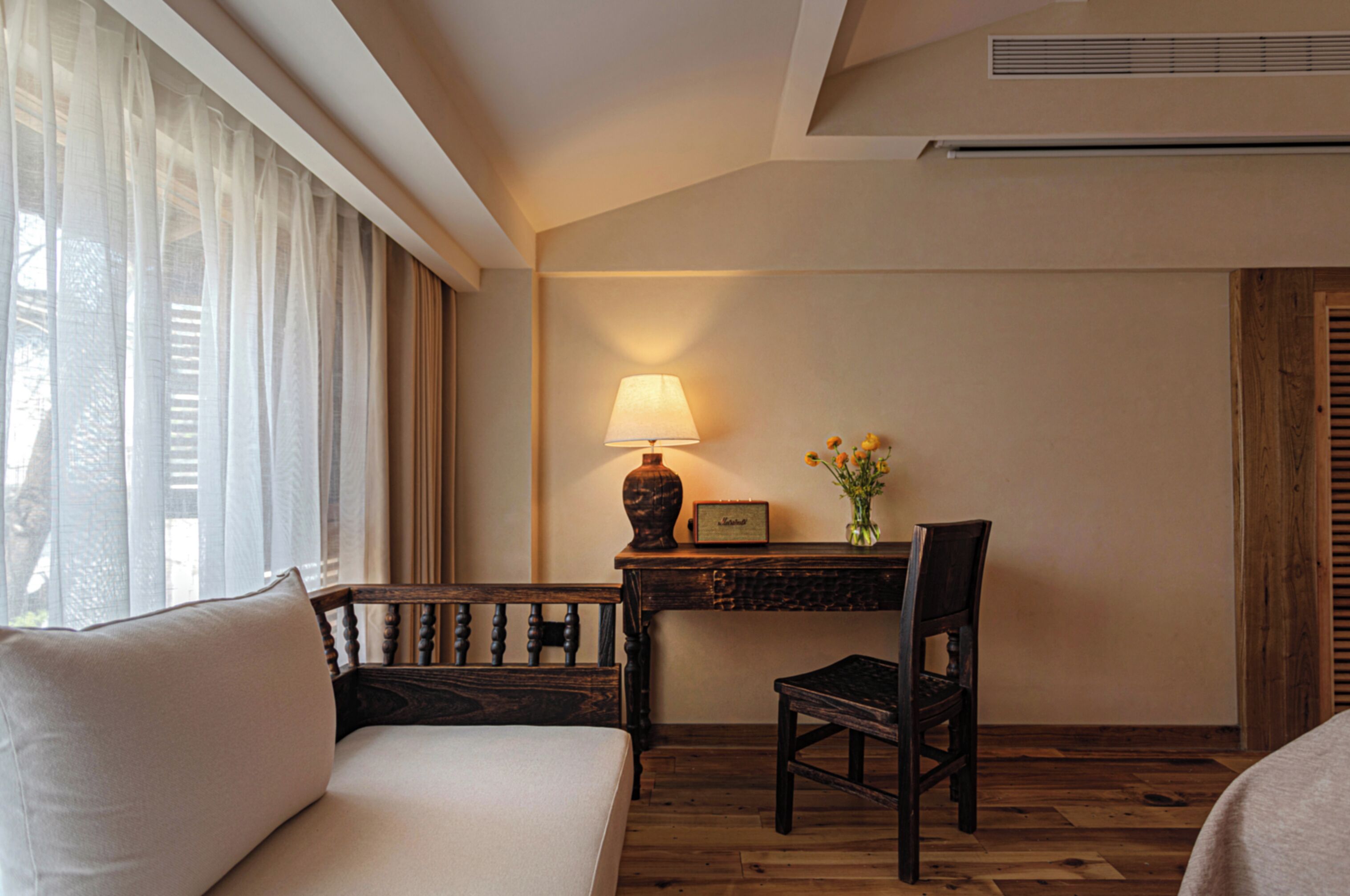 A hotel room with a bed, a desk with a lamp, a chair, and a window with sheer curtains.