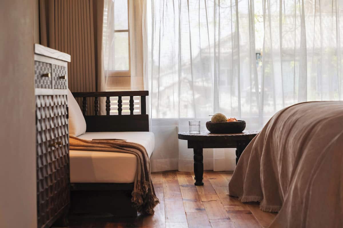 A bedroom with a bed, a bedside table with a bowl and glass, a window with sheer curtains, and a patterned headboard.
