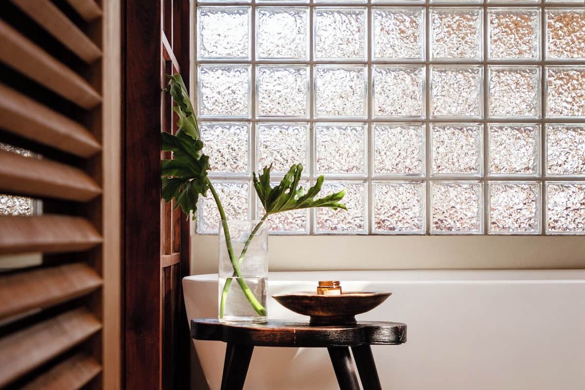 A room with a window featuring frosted glass panels, a wooden slatted partition, a small table with a vase and a candle, and a tiled floor.