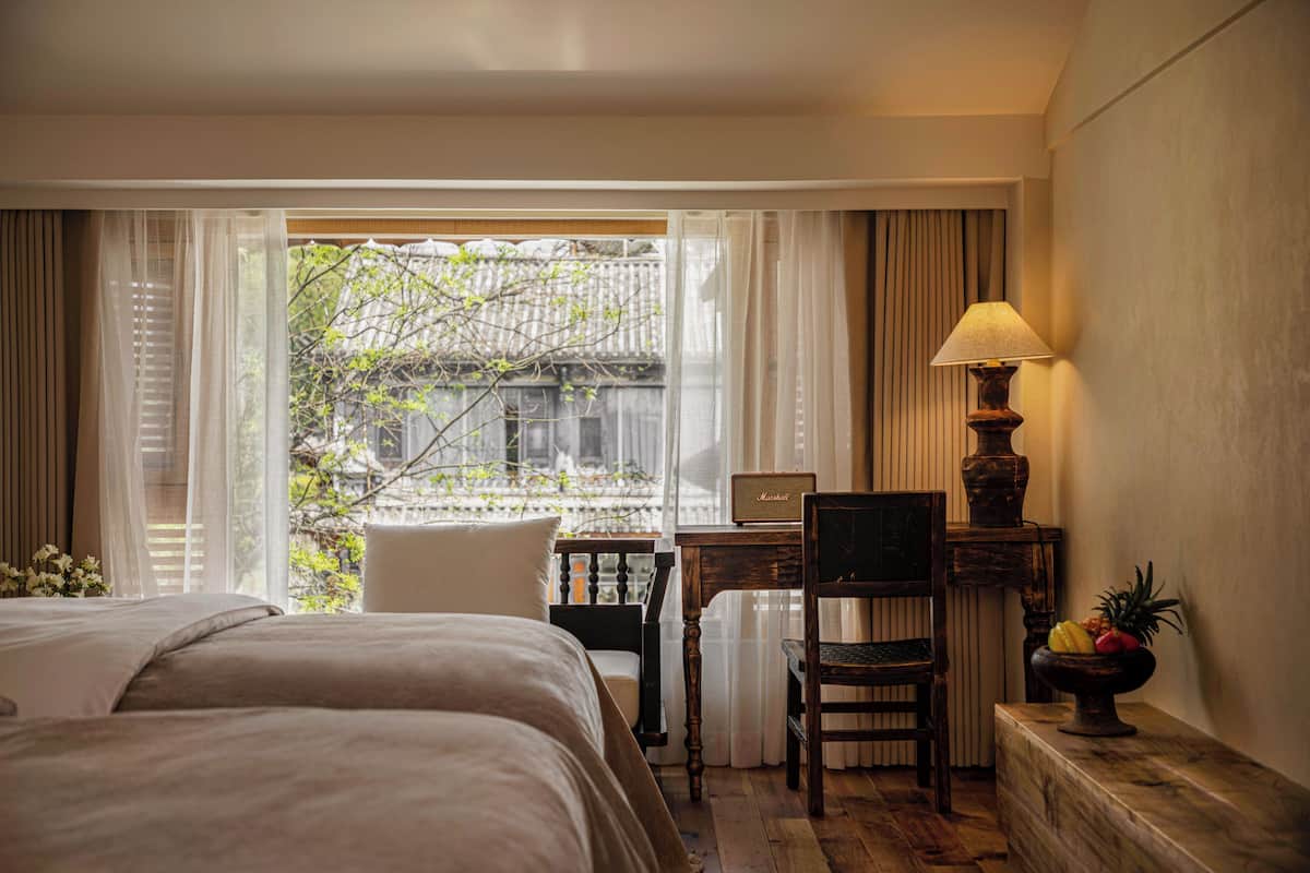 A hotel room with a bed, a desk, a chair, a lamp, and a fruit bowl.