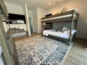 Uma cama beliche com um tapete estampado, um sofá com almofadas e uma TV fixada na parede.