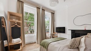 A bedroom with a bed, a wooden wardrobe, a television, a fireplace, and a window with curtains. - GuestReady - Modern Double En-Suite near Euston (London)