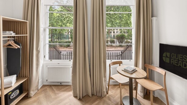 A room with a view of a park, a radiator, a wooden shelf with a radio, and a table with a chair. - GuestReady - Modern Double En-Suite near Euston (London)