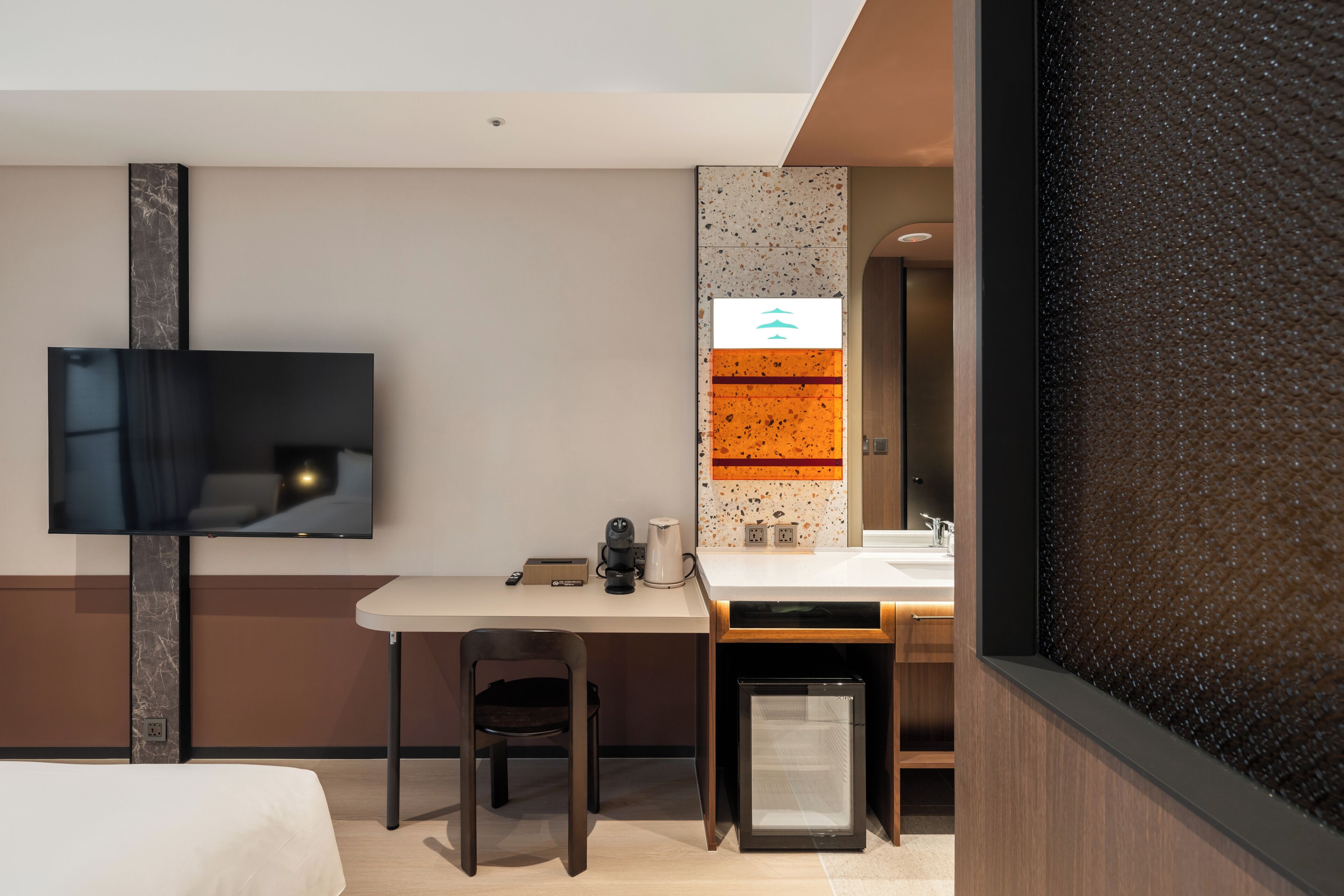 A hotel room with a flat-screen TV, a desk with a chair, a small refrigerator, a sink, and a mirror.