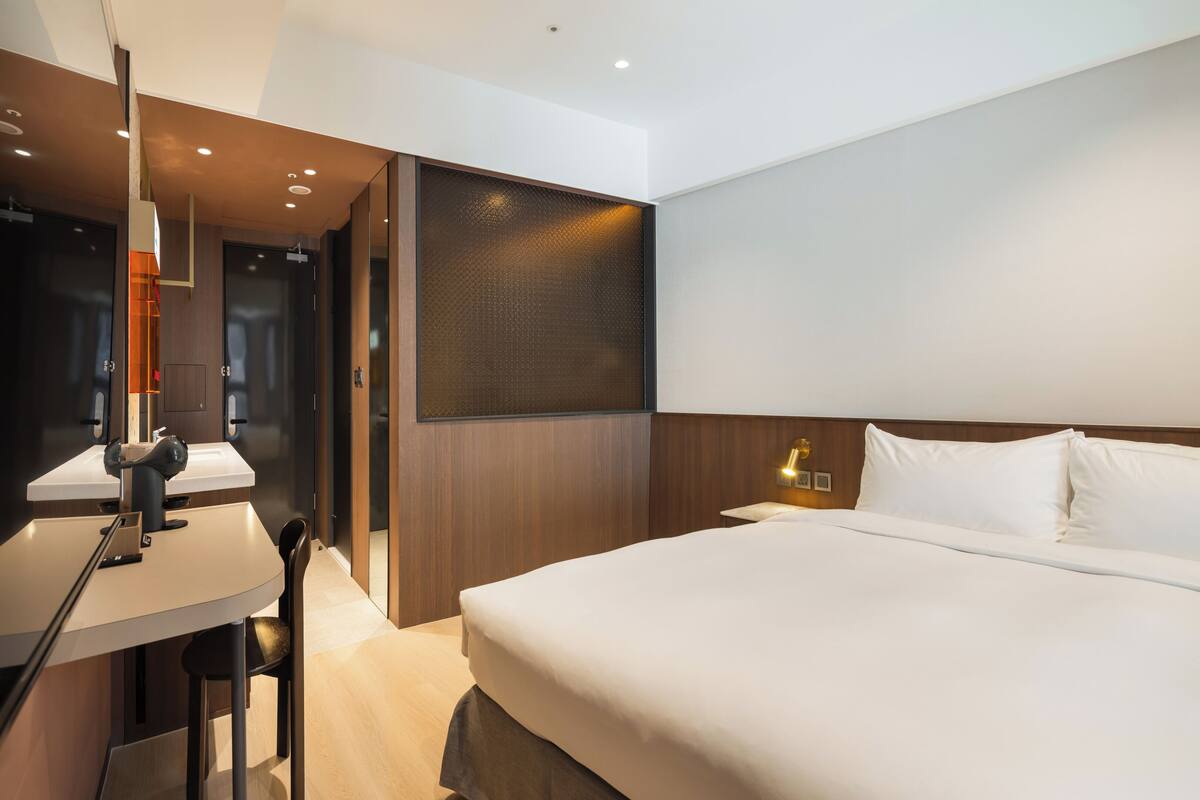 A modern hotel room with a large bed, a desk with a lamp, and a wall-mounted light fixture.
