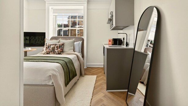 Une chambre à coucher avec un lit, un bureau, un miroir, une fenêtre et une vue sur les bâtiments à l’extérieur.