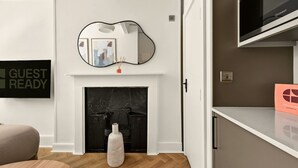 A modern living room with a fireplace, a mounted mirror, a TV, and a 'GUEST READY' sign. - GuestReady - Comfortable Double Room in London (London)