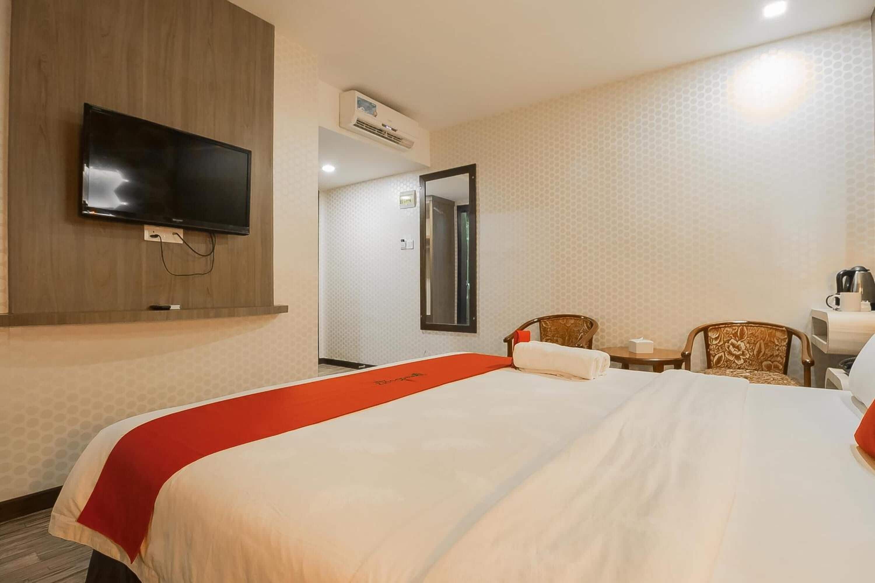 A hotel room with a bed, a TV mounted on the wall, a small table with a chair, and an air conditioning unit.