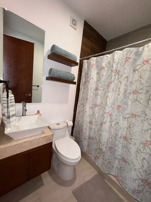 Ein Badezimmer mit weißem Waschbecken, Toilette, einer Duschvorhang mit Blumenmuster und zwei Ablagen über der Toilette.