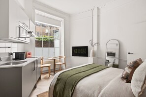 A modern, minimalist hotel room with a kitchenette, a large window, a bed with a green throw, and a 'GUEST READY' sign. - GuestReady - Modern Double En-Suite in Bloomsbury (London)