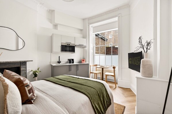 A modern bedroom with a bed, a fireplace, a kitchen area, and a view of a brick building through a large window. - GuestReady - Modern Double En-Suite in Bloomsbury (London)
