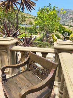 Un banco de madera con elaboradas esculturas, una barandilla de piedra y vistas a un jardín ajardinado con diversas plantas y una colina distante.
